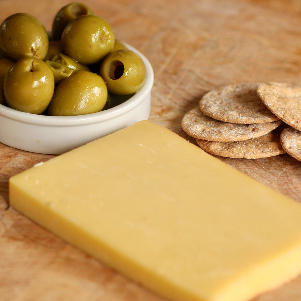 A block of smoked Cheddar cheese on a wooden surface, with a small white bowl of green olives and a stack of round brown crackers nearby.