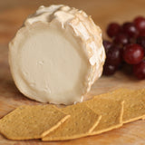 A round wheel of creamy goats cheese with a textured rind, shown on a wooden surface with crackers in front and red grapes in the background.