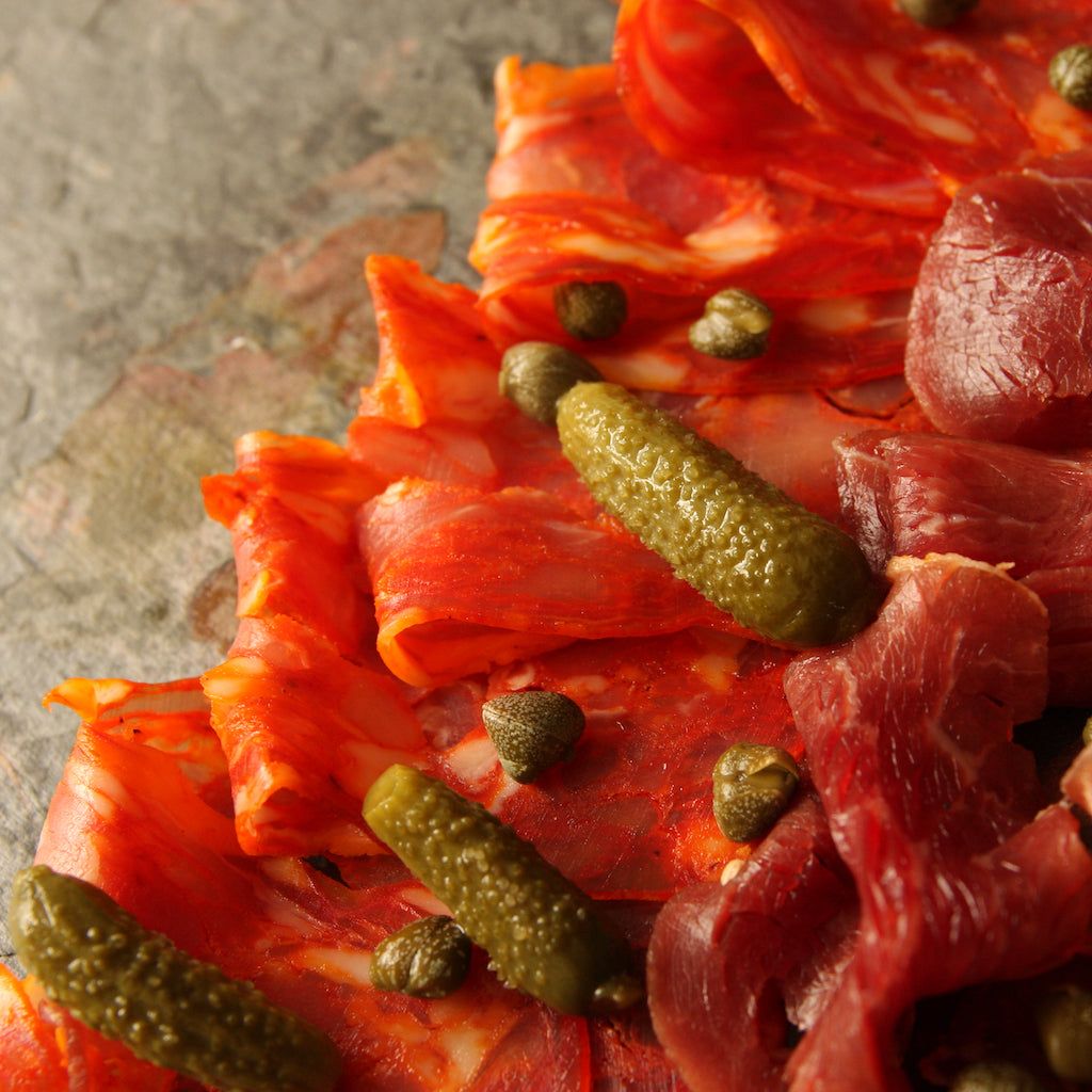 Smoked Spianata Calabra salami with other sliced smoked meats on slate with capers and gherkins