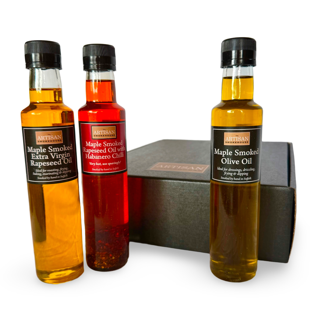 Three glass bottles of Artisan Smokehouse maple smoked oils arranged in front of a dark black gift box on a white background. The bottles are labelled Maple Smoked Extra Virgin Rapeseed Oil, Maple Smoked Habanero Chili Oil, and Maple Smoked Olive Oil.