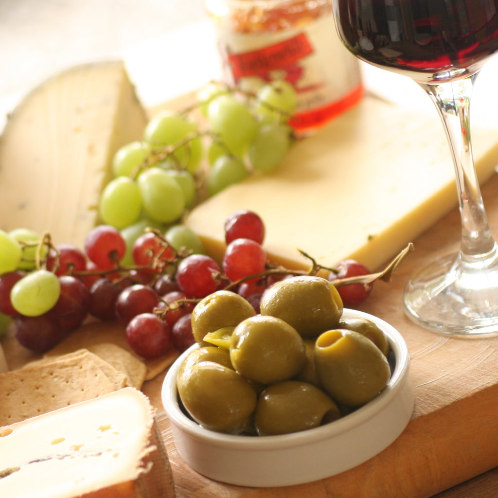A ramekin of The Artisan Smokehouse's maple smoked olives on wooden board with cheese, grapes and wine