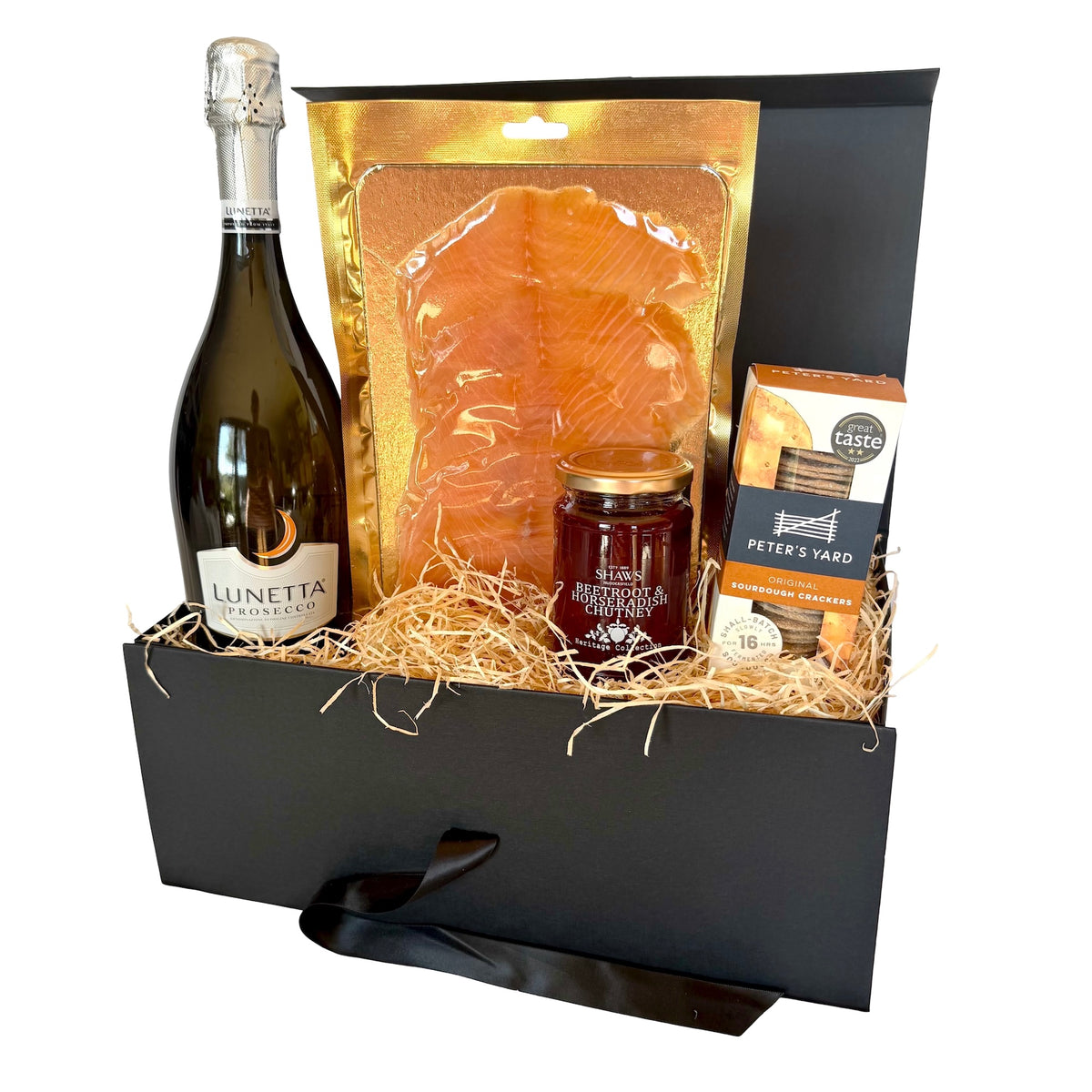A curated gift basket in a black box with straw filler, featuring a bottle of Lunetta Prosecco, a large bag of smoked salmon, a jar of preserves, and a box of Peter's Yard crackers, set against a plain white background.