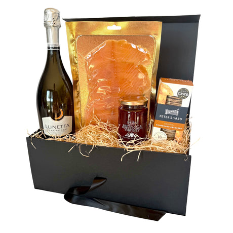 A curated gift basket in a black box with straw filler, featuring a bottle of Lunetta Prosecco, a large bag of smoked salmon, a jar of preserves, and a box of Peter's Yard crackers, set against a plain white background.