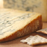 A close-up of a wedge of smoked Stilton cheese on a wooden surface with crackers in the foreground and a blurred cheese block in the background.