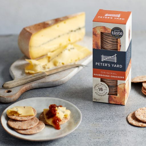 A box of Peter's Yard Original Sourdough Crackers stands on a gray surface beside a cutting board with a large wheel of cheese, a knife, and a plate with crackers and cheese slices. 