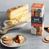 A box of Peter's Yard Original Sourdough Crackers stands on a gray surface beside a cutting board with a large wheel of cheese, a knife, and a plate with crackers and cheese slices. 