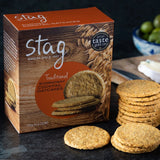 A box of Stag Traditional Stornoway Cocktail Oatcakes with several round oatcake slices stacked and placed in front on a dark tabletop, with a blurred bowl of green olives in the background.
