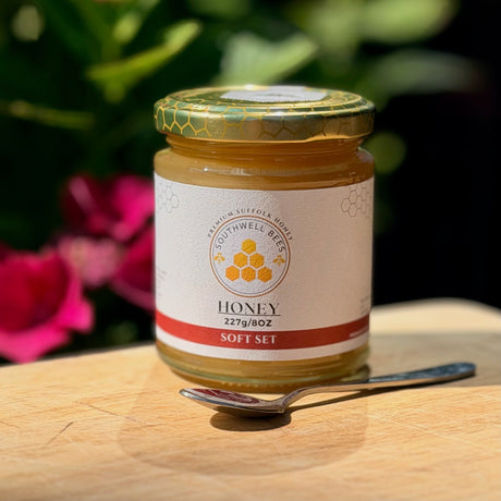 A jar of Southwell Bees Honey on a wooden surface with a spoon, set against a blurred background of green leaves and pink flowers.