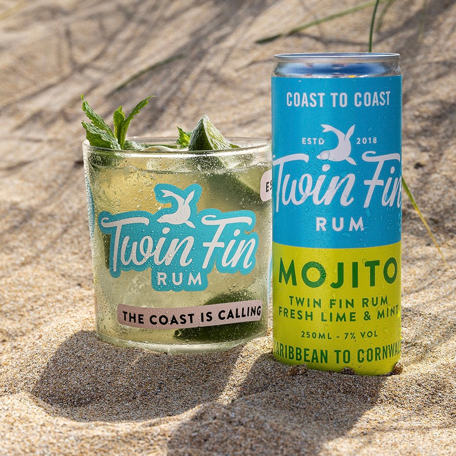 A can of Twin Fin Rum Mojito and a glass of the same mojito cocktail sit on a sandy beach.