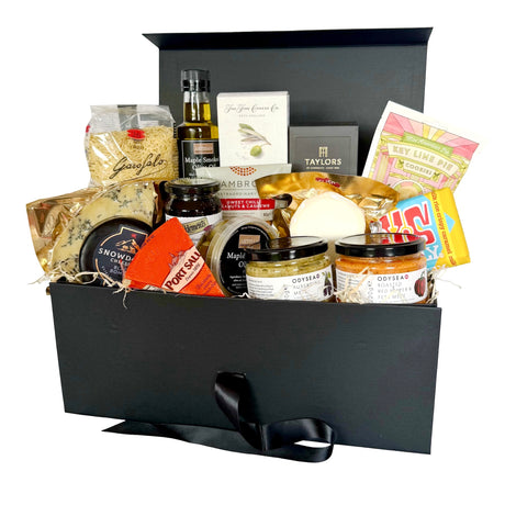 A vegetarian hamper filled with an assortment of food items, including cheeses, spreads, olives and nuts, arranged inside an open black gift box with a black ribbon handle on a white background.