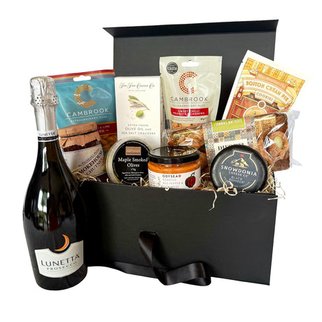 A gourmet gift hamper in a black box, featuring a bottle of Lunetta Prosecco, Cambrook corn, a jar of Odysea meze , a round cheese from Snowdonia, and other packaged snacks and sweets, arranged in a black gift box.