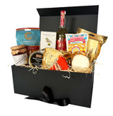 A gourmet vegetarian picnic hamper in an open black box with a black ribbon bow. The box is filled with various cheeses, crackers, a bottle of peach Bellini, and packaged snacks on a white background.