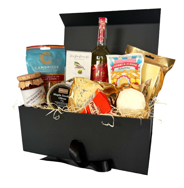 A gourmet vegetarian picnic hamper in an open black box with a black ribbon bow. The box is filled with various cheeses, crackers, a bottle of peach Bellini, and packaged snacks on a white background.