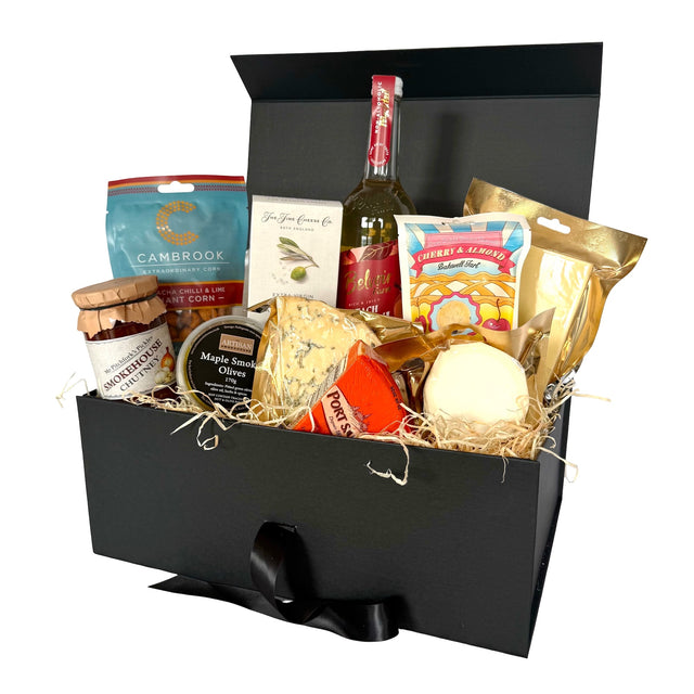 A gourmet vegetarian picnic hamper in an open black box with a black ribbon bow. The box is filled with various cheeses, crackers, a bottle of peach Bellini, and packaged snacks on a white background.